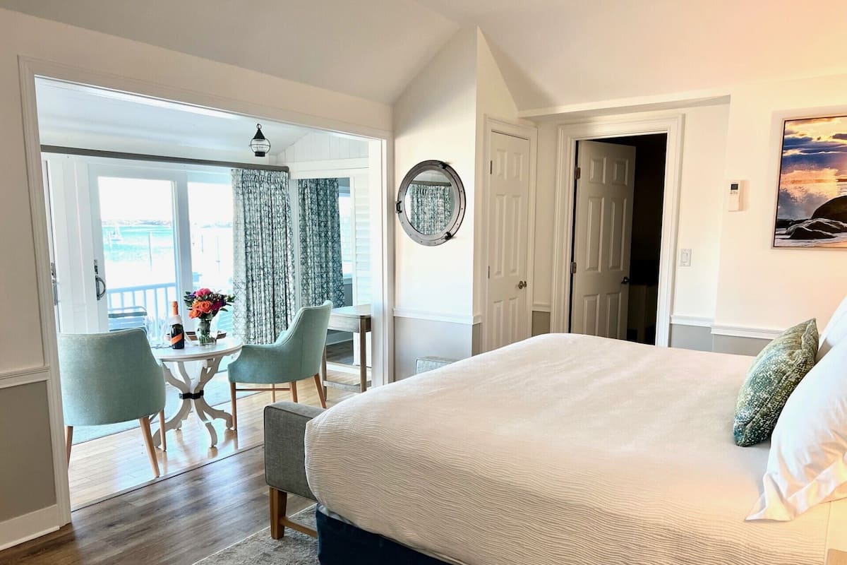 Un dormitorio con una cama grande, un comedor con mesa y sillas, y vistas a un balcón exterior a través de un ventanal.