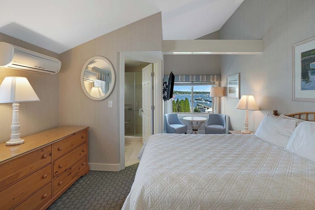 Habitación de hotel con cama, cómoda, espejo redondo, dos sillas, una mesita y vistas al exterior a través de una ventana.