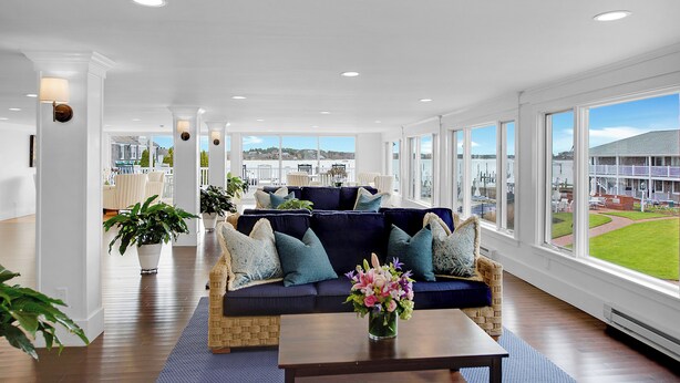 A spacious room with a dark blue sofa, white walls, and large windows offering a view of a building and a grassy area. - The Harborside Inn (Edgartown)