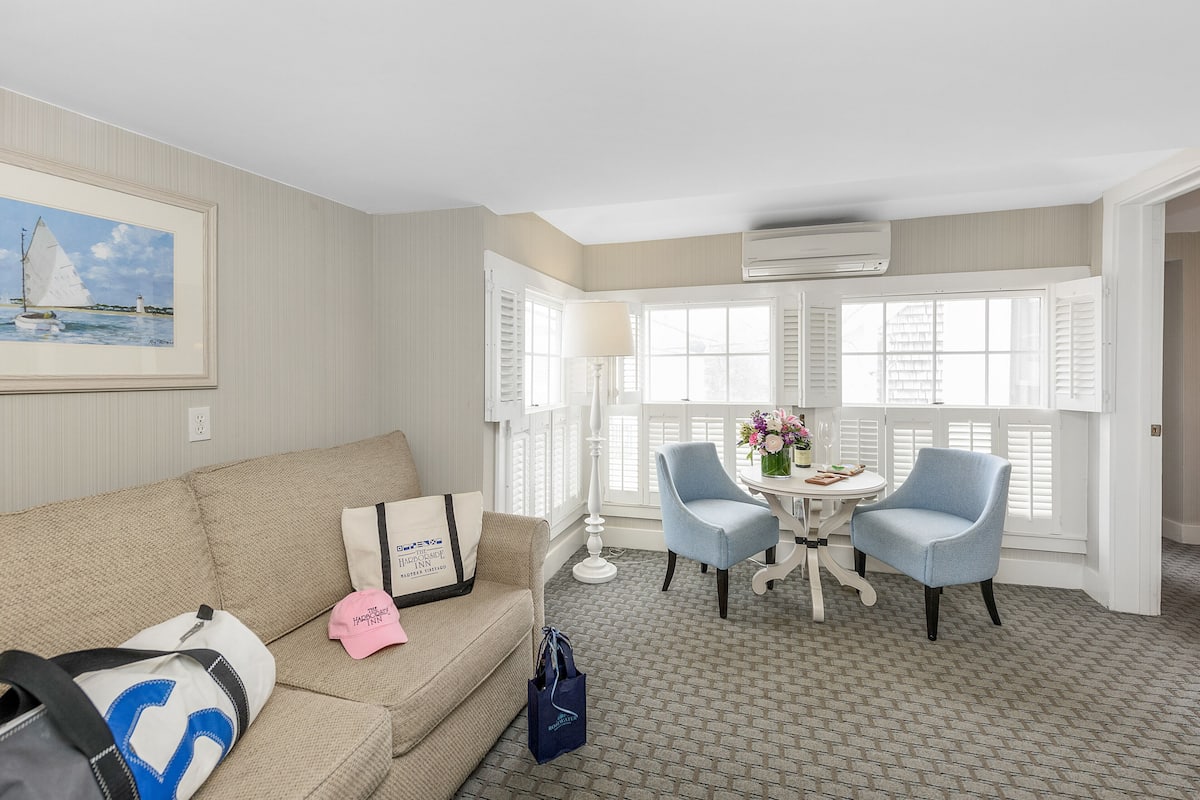 Habitación de hotel con un sofá color beige, dos sillones, una mesita con un florero y una ventana con contraventanas.