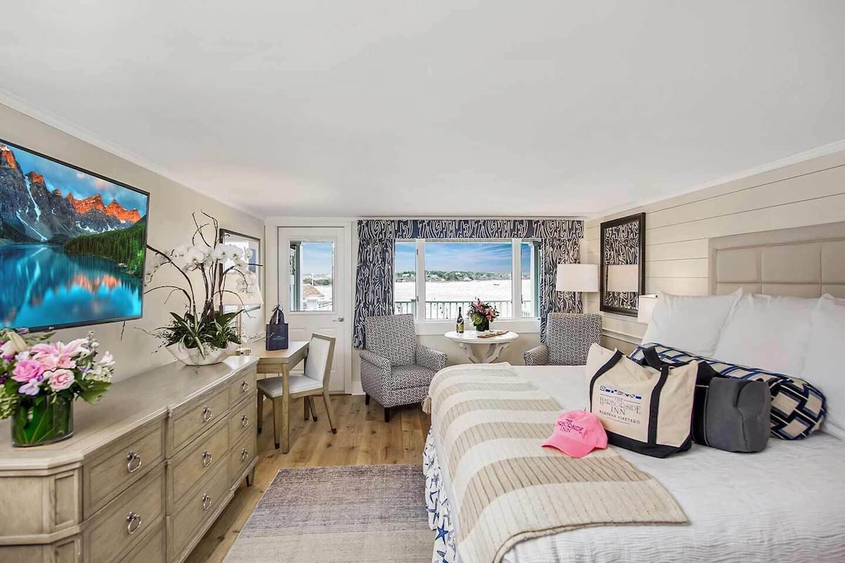 Habitación de hotel con cama, escritorio, silla, pequeña mesa con botella y flores, televisor y vista a la playa.