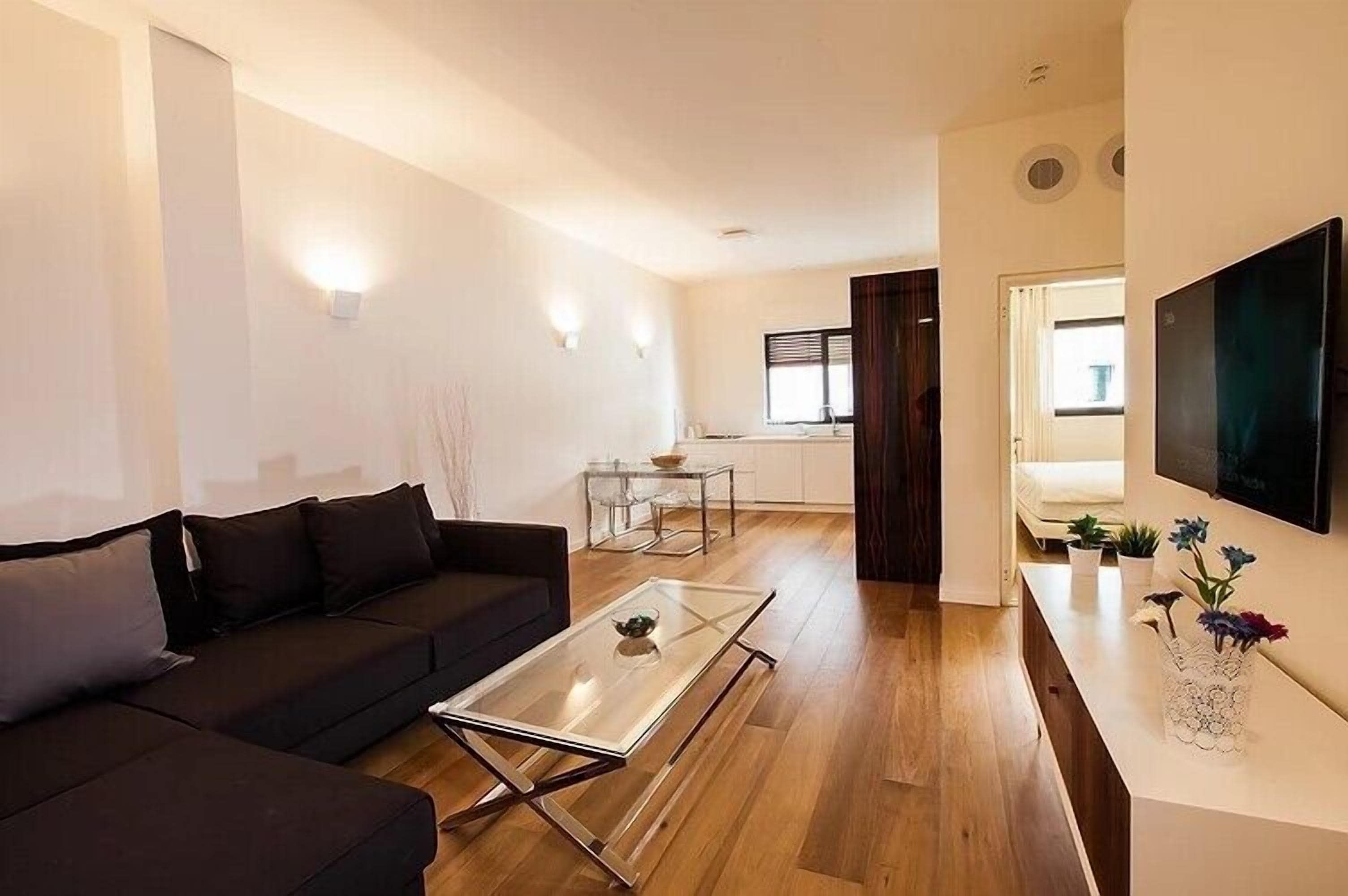 A modern living room with a black sofa, a glass coffee table, and a TV on the wall.