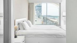 A hotel room with a large bed, a nightstand, a lamp, and a view of a cityscape and beach. - Tel Aviv Home (Tel Aviv)