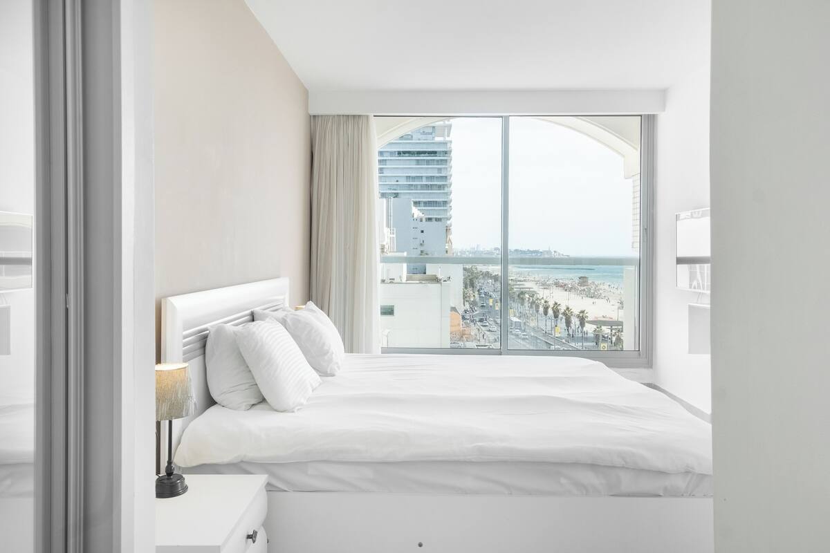 Una habitación de hotel con una cama grande, una mesita de noche, una lámpara y vistas a la ciudad y a la playa.