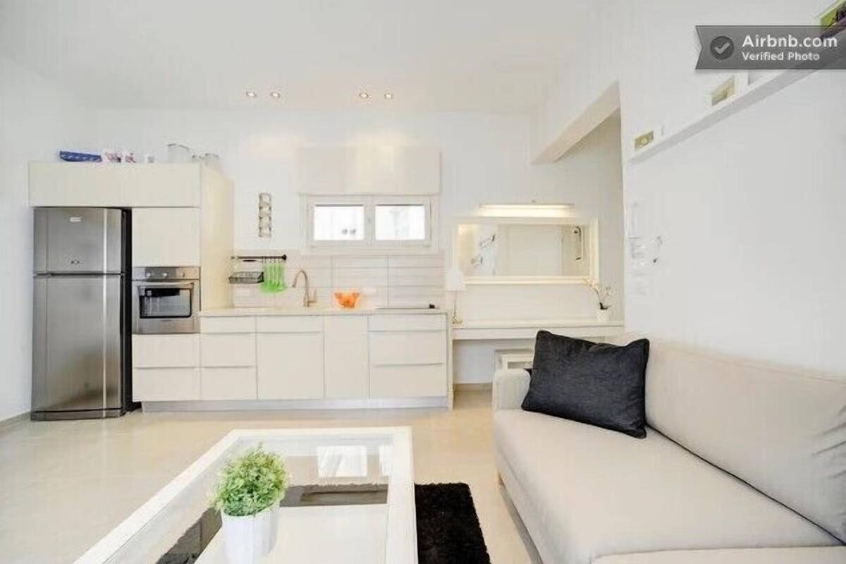 Una cocina moderna con armarios blancos, horno empotrado y refrigerador. Hay un comedor con una mesa blanca y un tapete negro.