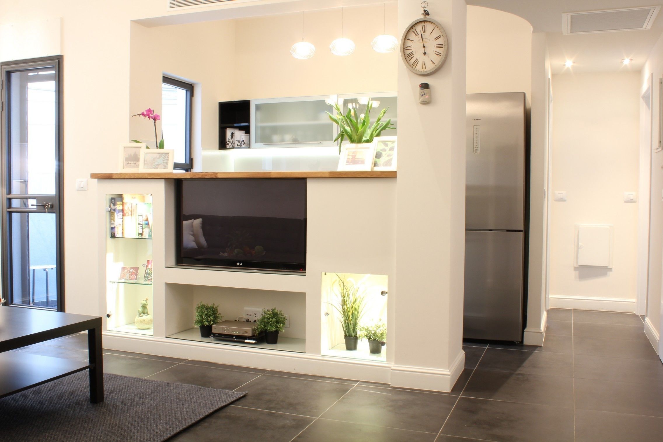 A modern living room with a flat-screen TV, built-in shelves, a clock, and a refrigerator.
