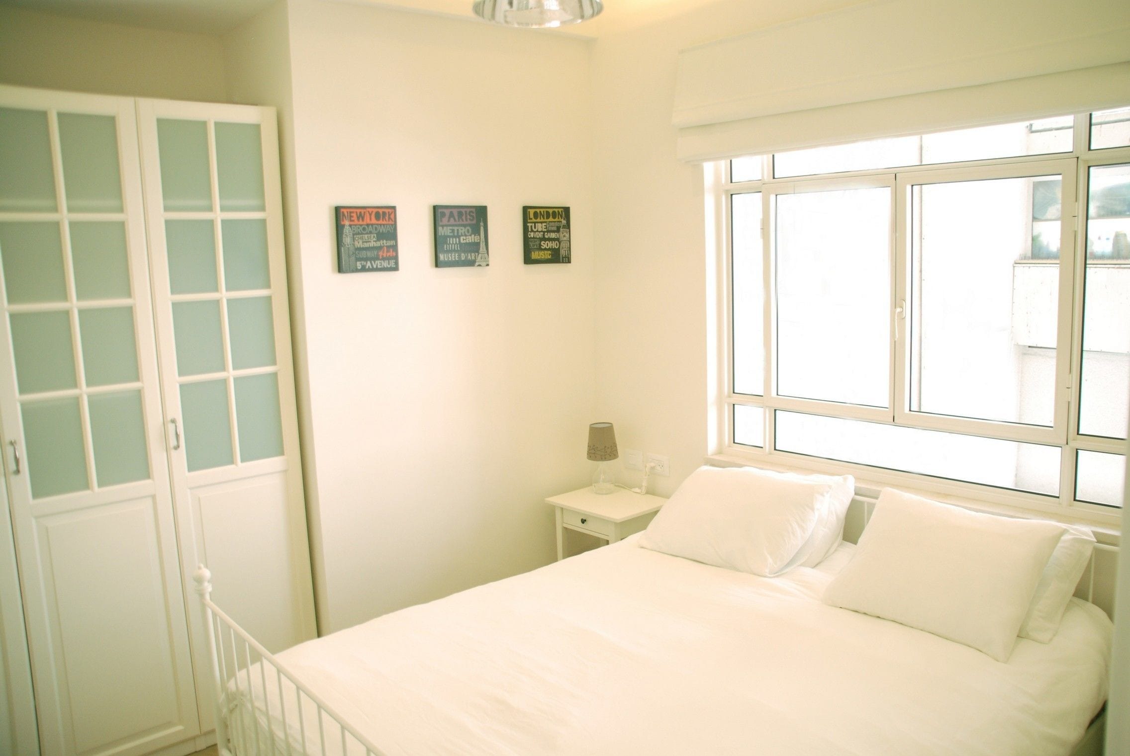 A bedroom with a bed, a nightstand, a lamp, and a window. There are three posters on the wall.
