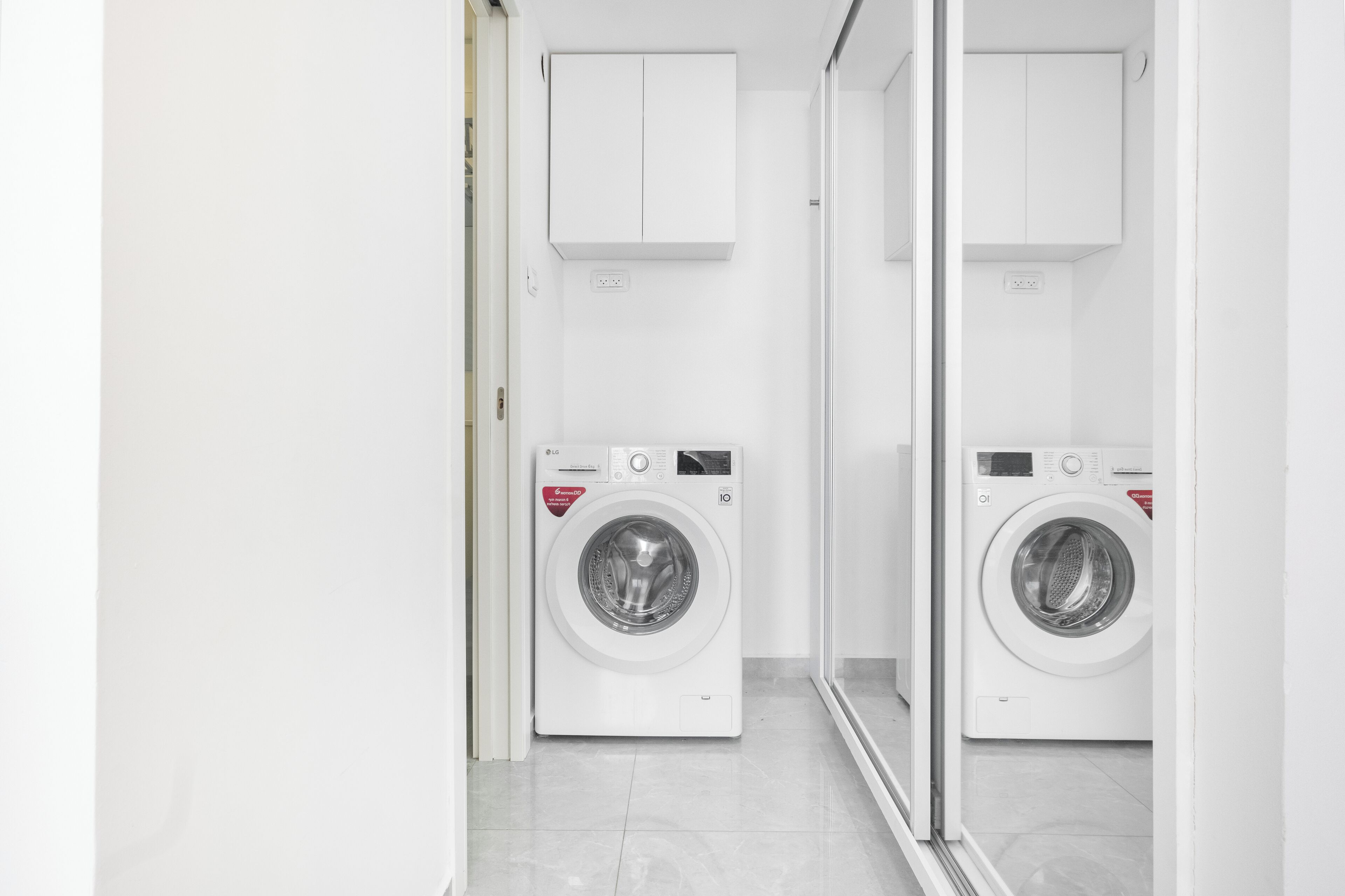 A clean, modern laundry room with two white washing machines, a mirror, and a white cabinet above the machines.