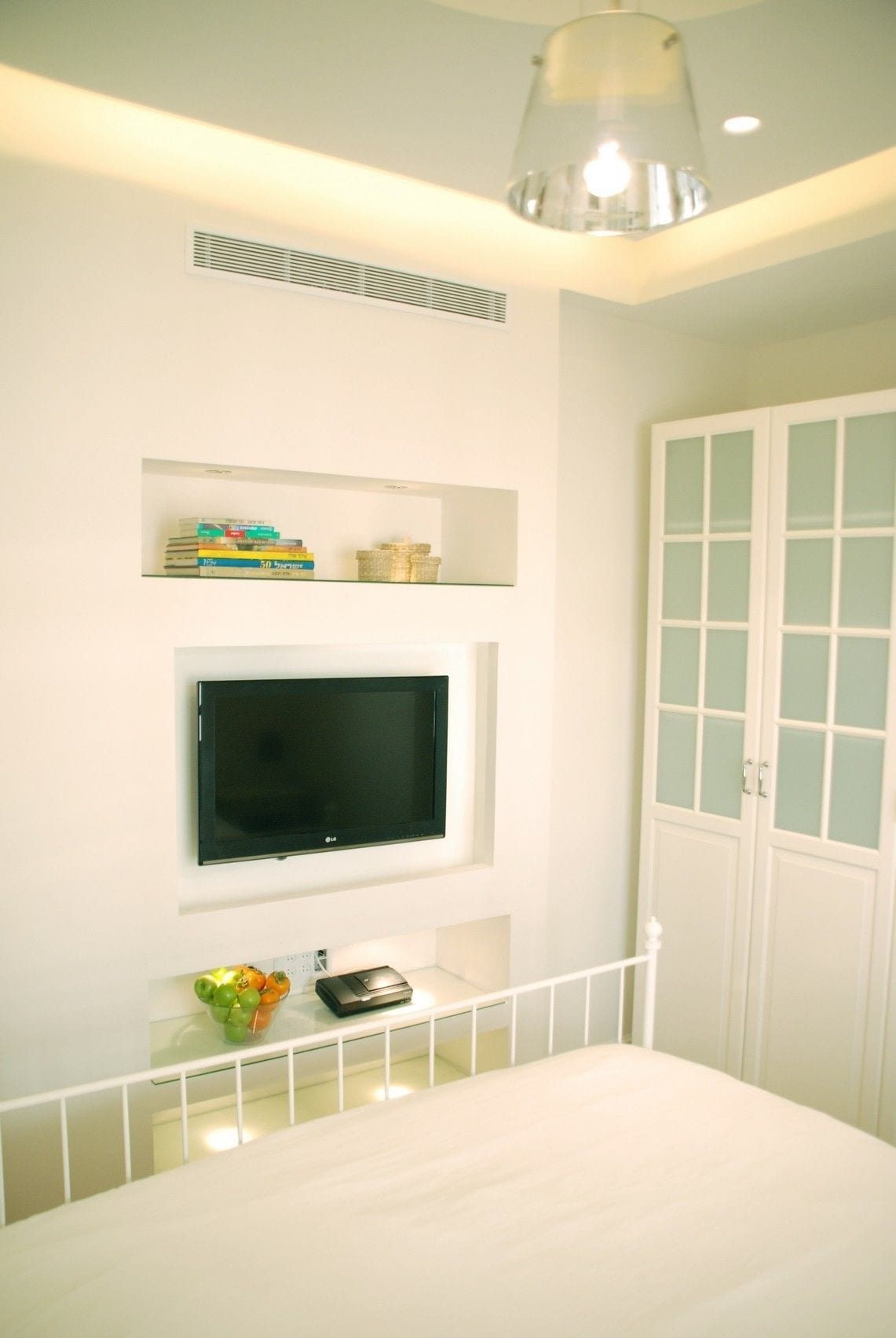 A bedroom with a bed, a TV mounted on the wall, a small shelf with books and decorative items, and a white wardrobe with frosted glass doors.