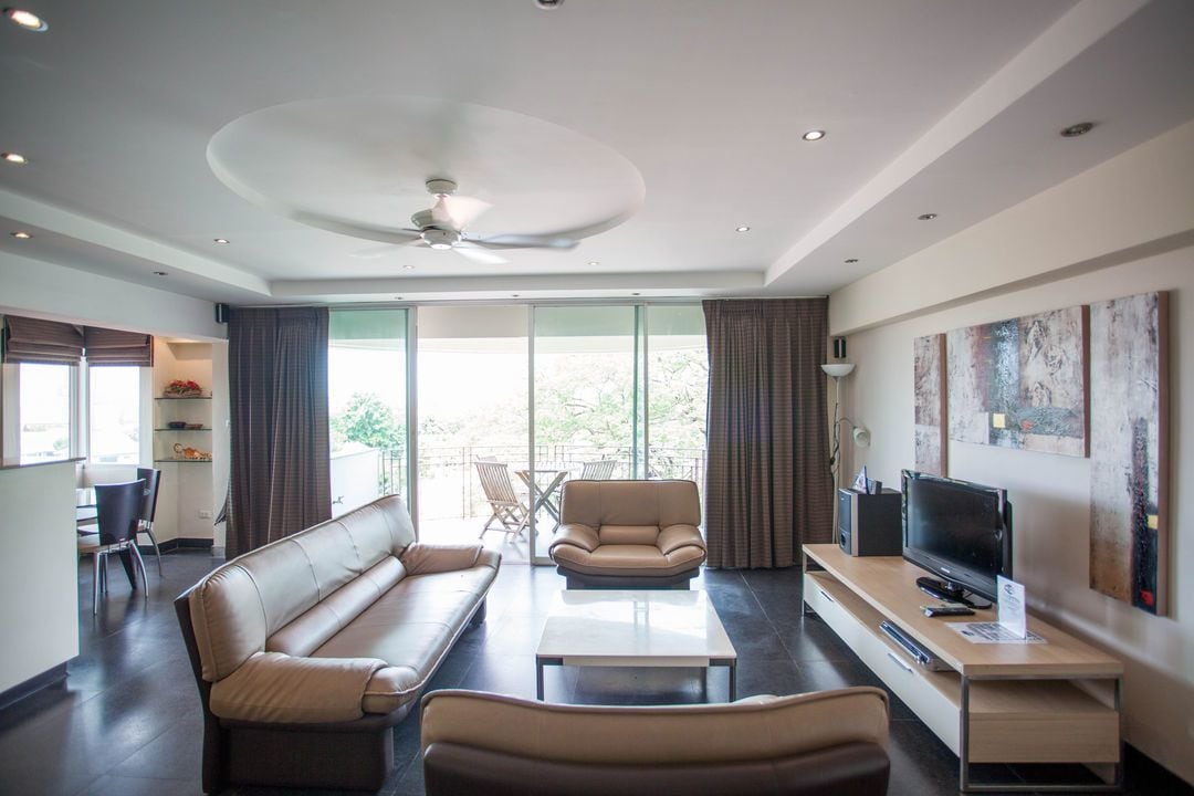 A modern living room with a sofa, armchairs, a coffee table, and a TV. There is a balcony with a table and chairs.