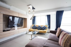 Een moderne woonkamer met een flatscreen televisie, een gele bank met opvallende kussens, een bruine bank met gestreepte kussens en een glazen salontafel.