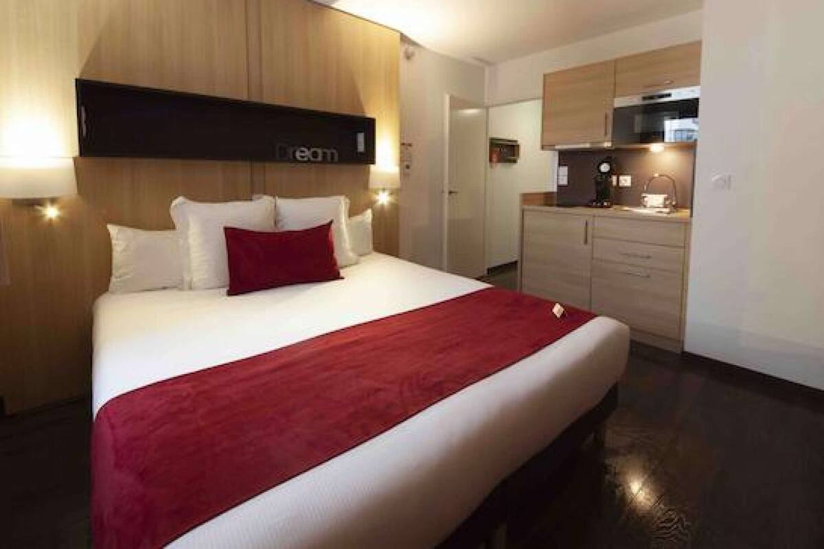 Habitación de hotel con una cama grande, cabecera de madera, manta roja y una kitchenette con microondas y un mueble pequeño.