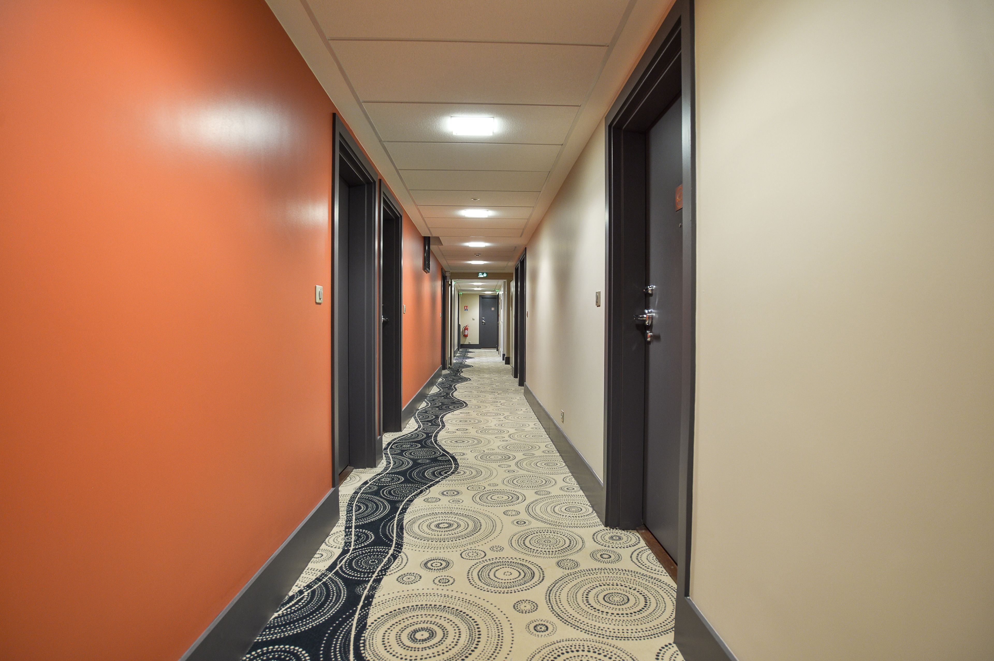 A hotel hallway with a patterned carpet, orange walls, and doors on both sides.