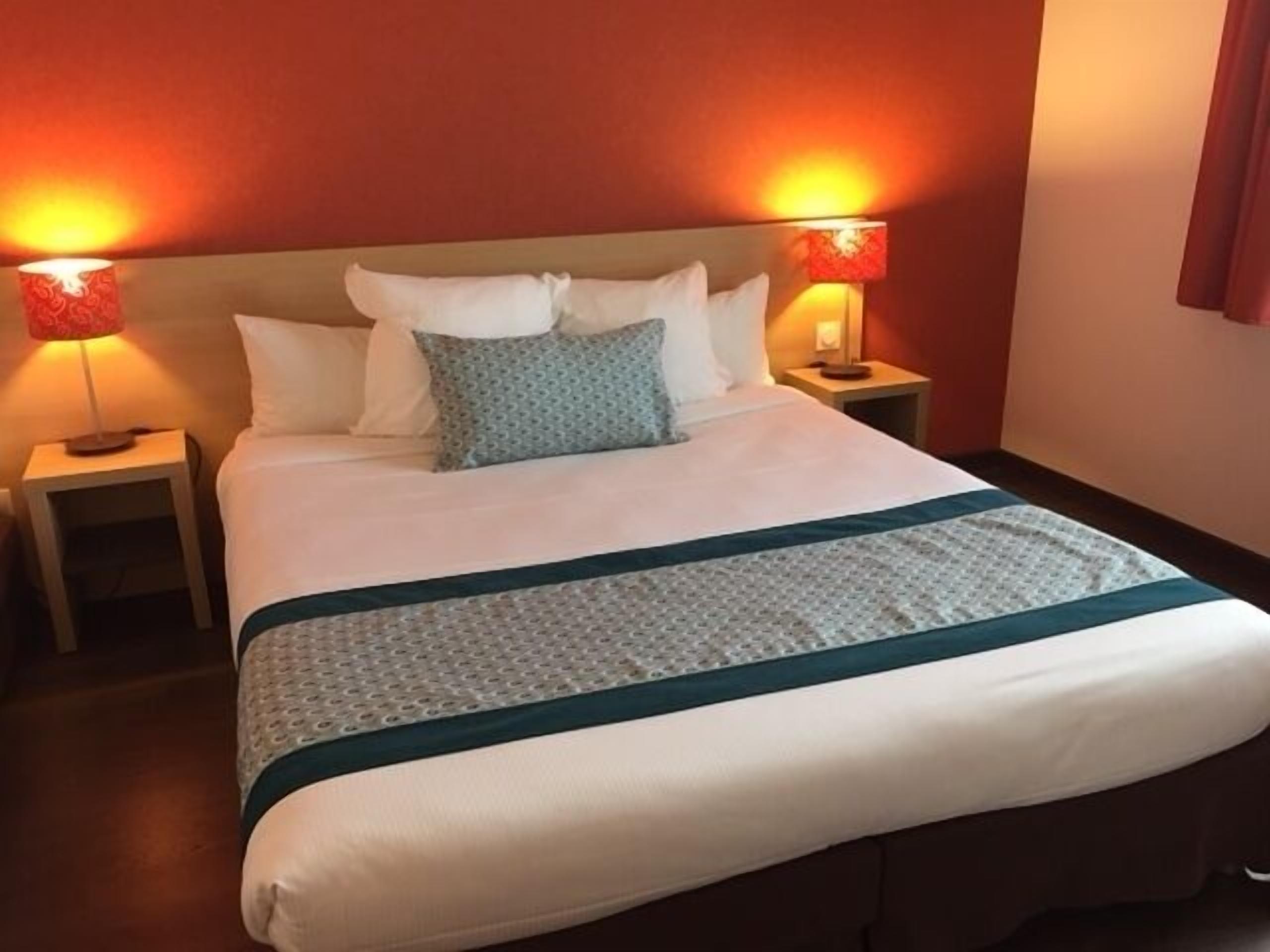 Un lit bien fait, avec du linge de lit blanc, un coussin turquoise et un plaid à motifs. De chaque côté du lit, on trouve deux lampes de chevet avec des abat-jour rouges.