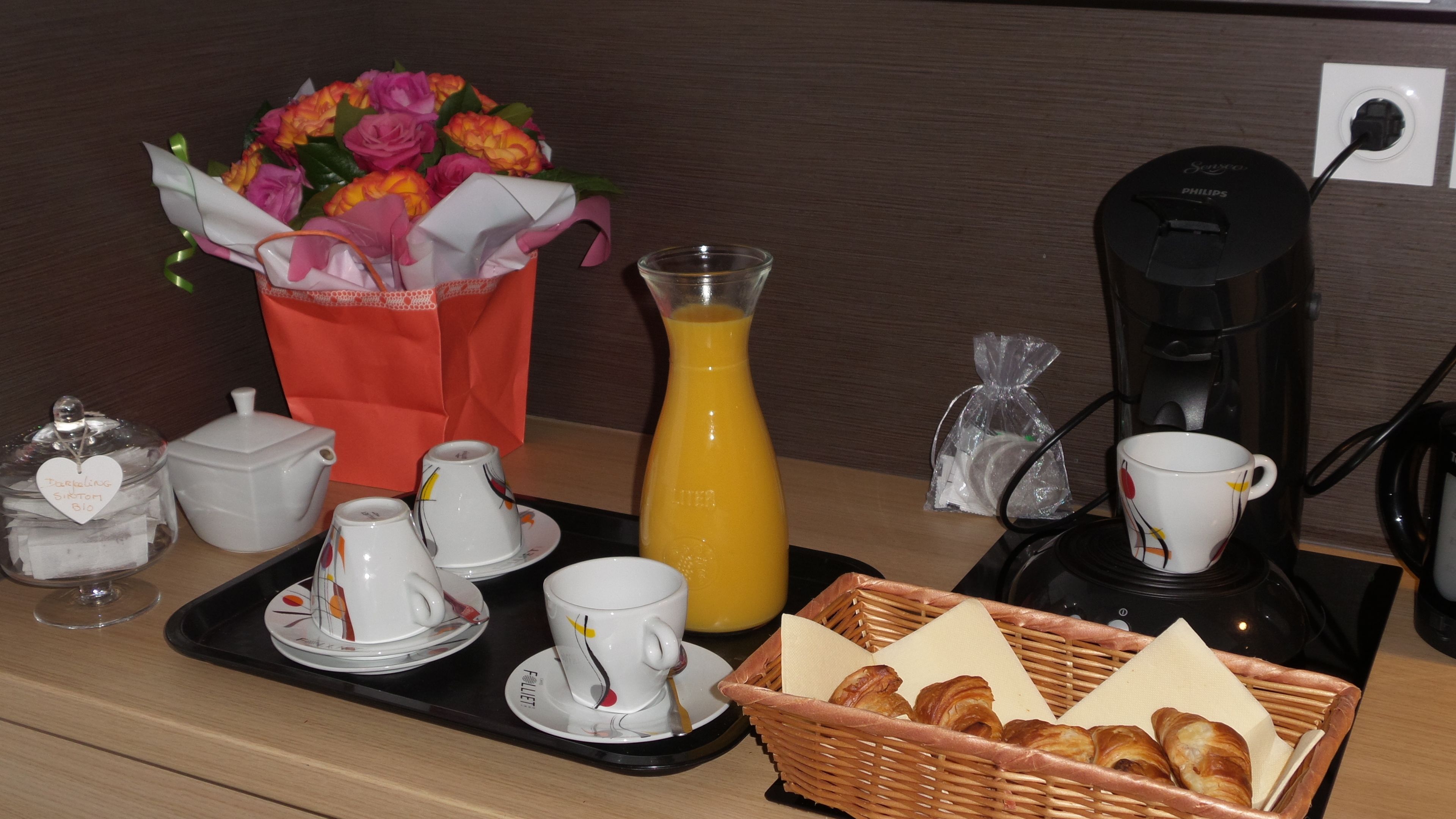 A breakfast setup with coffee maker, tea set, juice, pastries, and a bouquet of flowers.