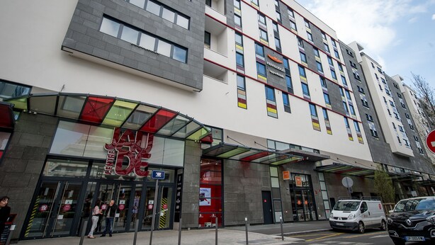 Un hotel de varios pisos con diseño moderno, que destaca por una entrada con puertas de vidrio y un toldo rojo.