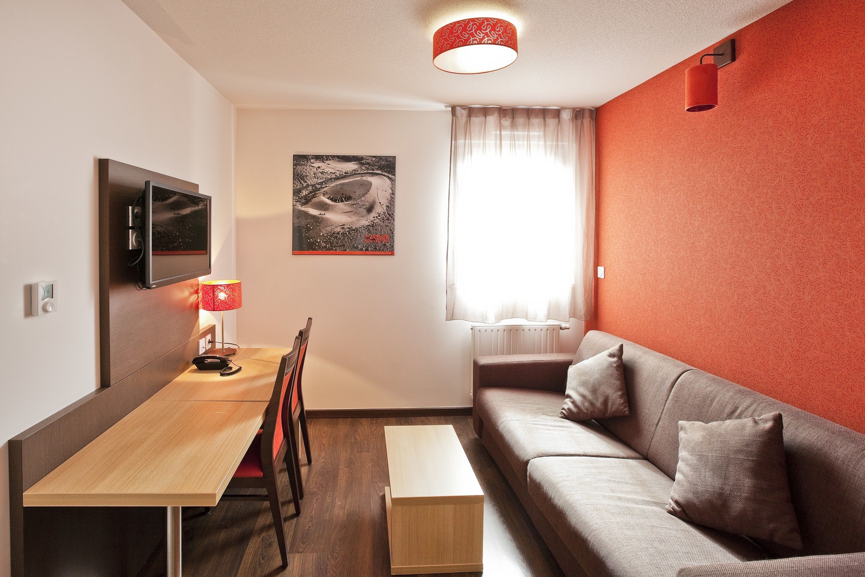A modern living room with a wooden dining table, a sofa, a TV mounted on the wall, and a window with curtains.