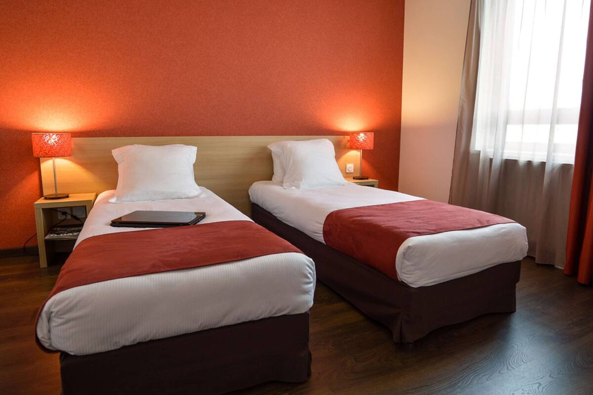 Habitación de hotel con dos camas individuales, cada una con mesita de noche y lámpara, suelo de madera y una ventana con cortinas.