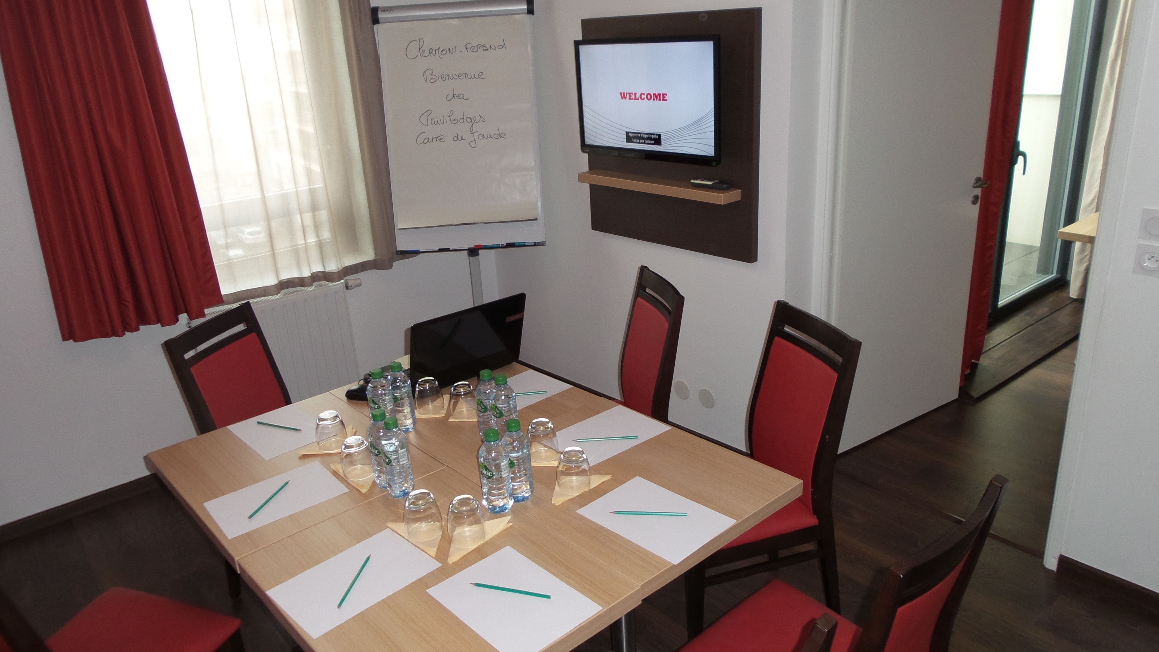 Una sala de conferencias con una mesa de madera, sillas rojas, una pizarra y una pantalla de televisión que muestra la palabra 'WELCOME'.