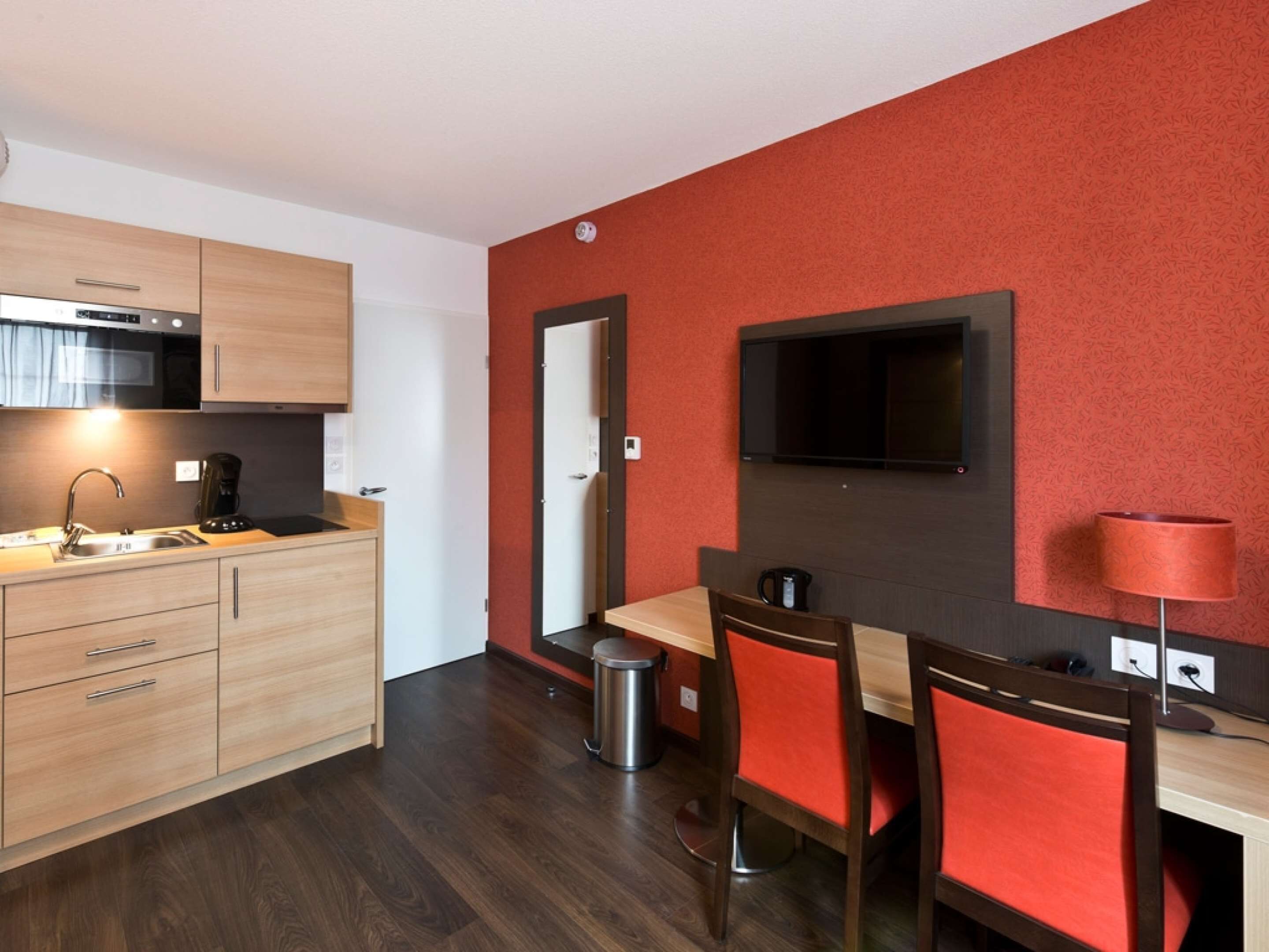 Une cuisine compacte comprenant un évier, une cuisinière et des placards, un coin repas avec une table et des chaises, et un mur d’accent de couleur rouge.