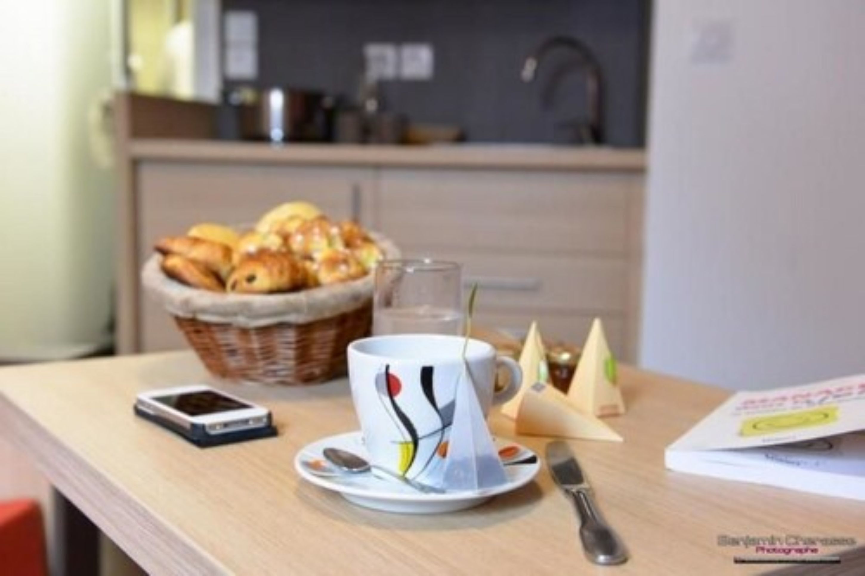 A breakfast setup with a basket of croissants, a cup with a colorful design, a glass of water, a smartphone, a knife, and a napkin cone.