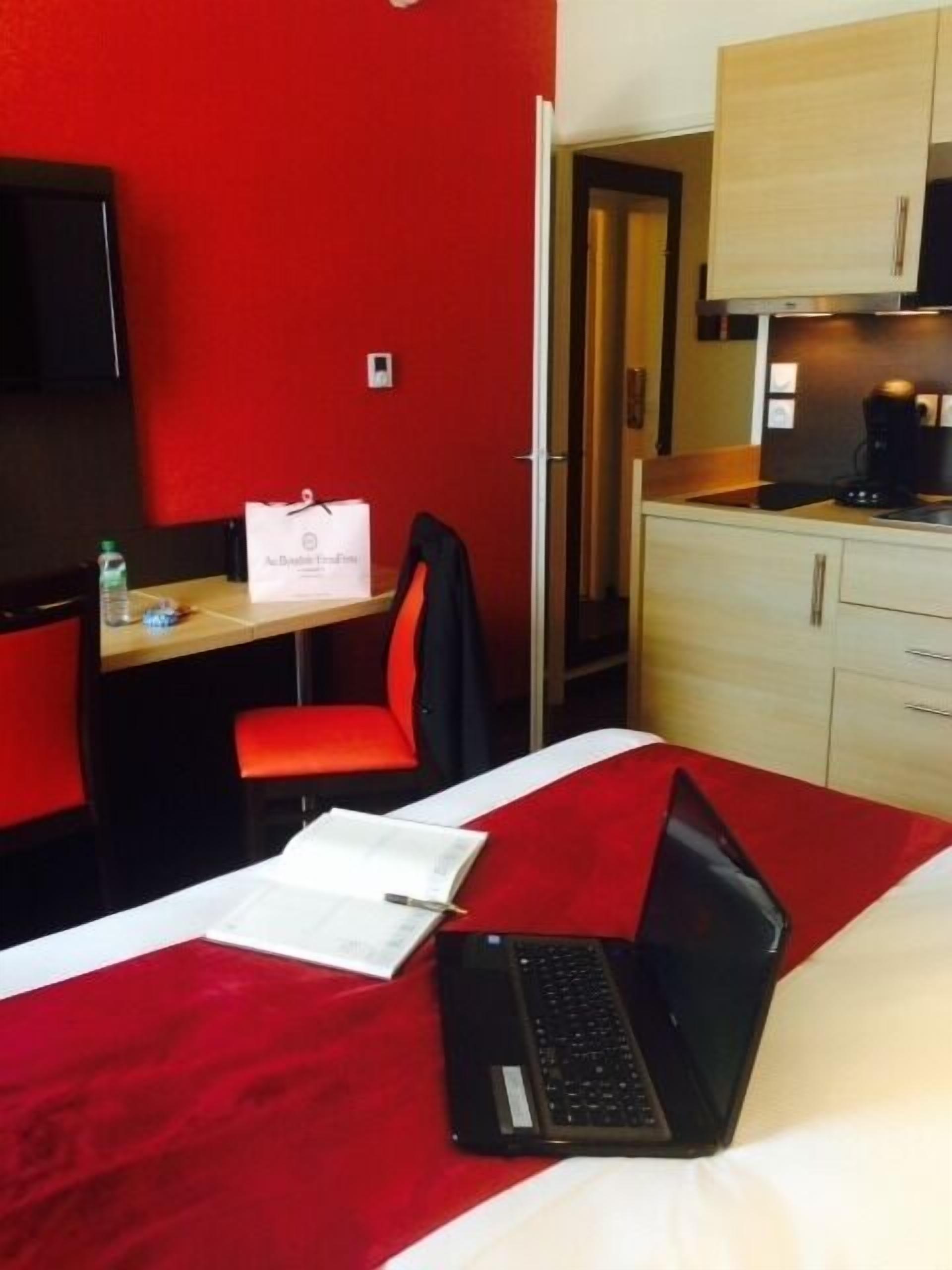 A hotel room with a red carpet, a desk with a laptop, a chair, a bed, a minibar, and a television.