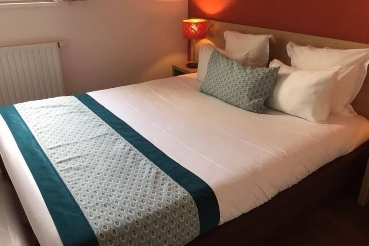 Una cama bien hecha con una colcha blanca y azul turquesa, una lámpara roja en la mesita de noche y una pared roja al fondo.