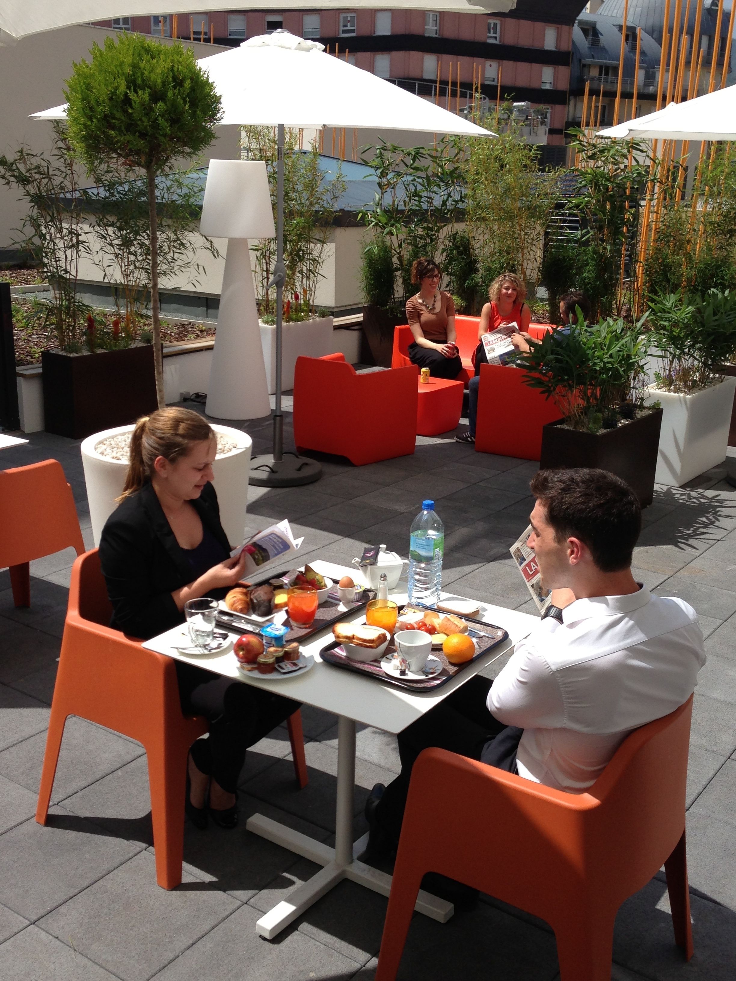 An outdoor dining area with orange chairs and tables, surrounded by potted plants and umbrellas.