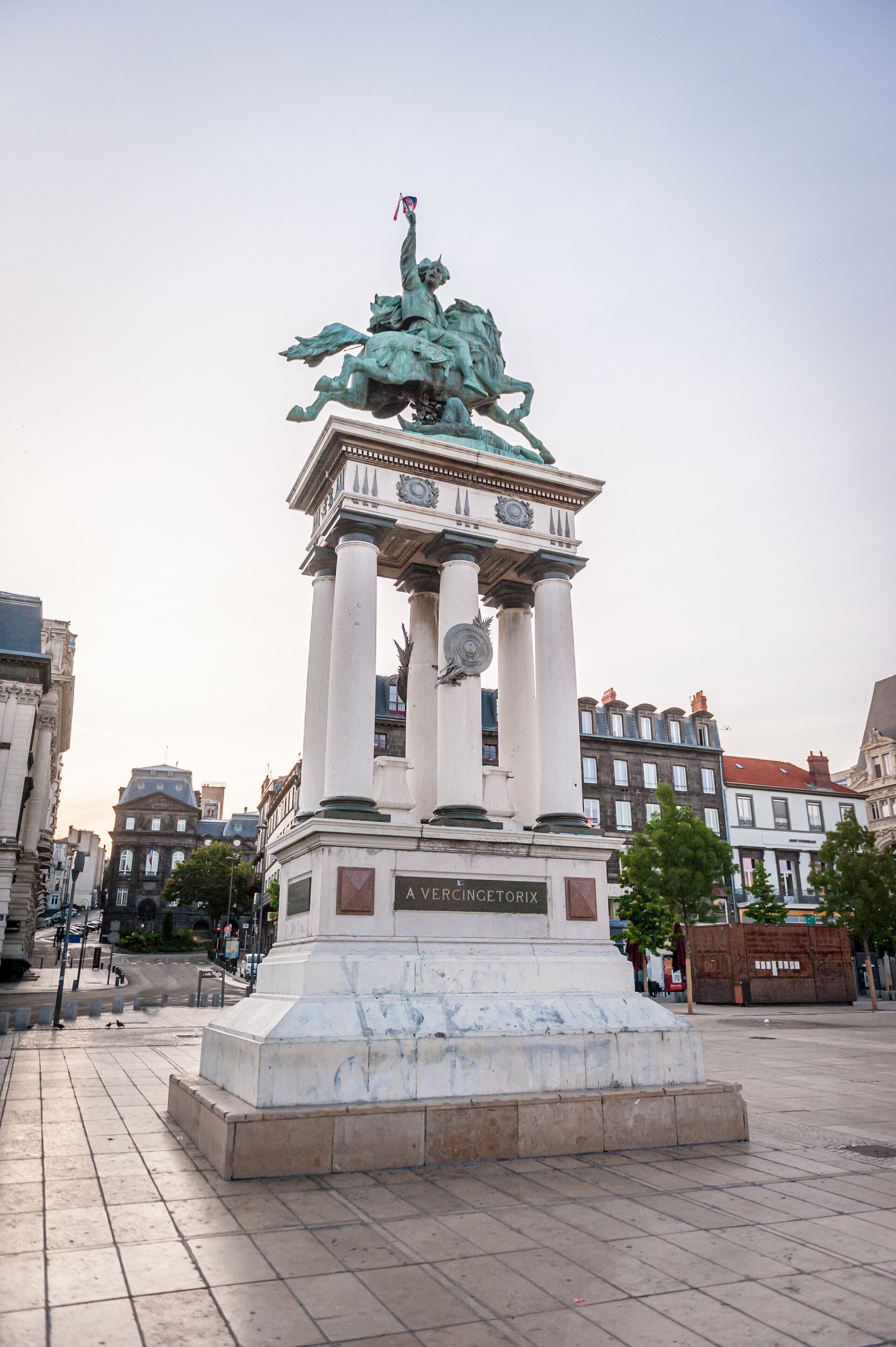 Un monumento con una estatua en la parte superior, rodeado por una plaza y edificios al fondo.