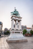 Un monumento con una estatua en la parte superior, rodeado por una plaza y edificios al fondo.