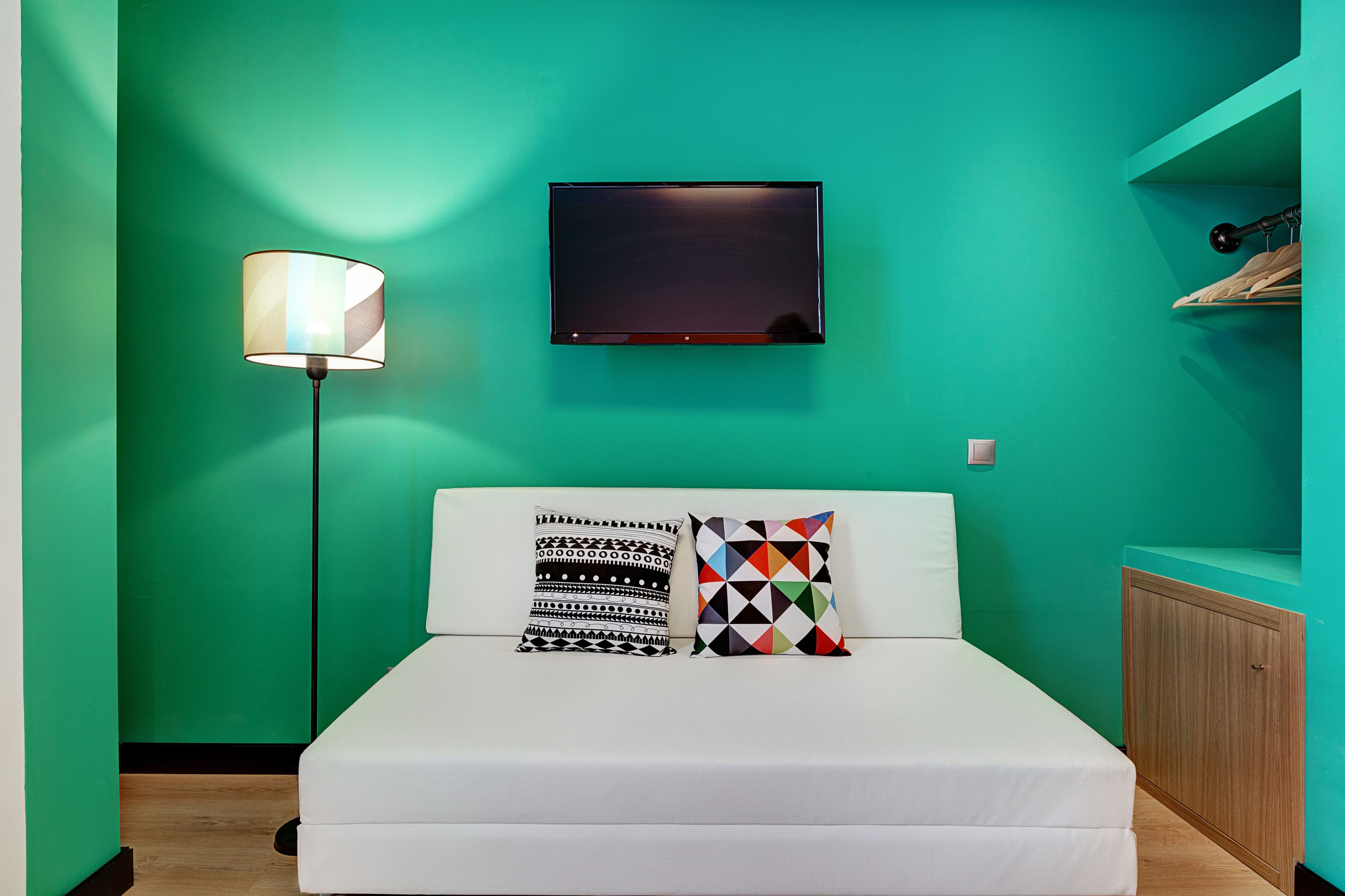 Ein Schlafzimmer mit einem weißen Bett, zwei Kissen, einer Stehlampe, einem an der Wand befestigten Fernseher und einem Holzschrank.