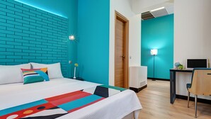 Une chambre d’hôtel moderne avec un mur d’accent de couleur turquoise, un lit soigneusement fait avec un couvre-lit aux motifs géométriques et colorés, un bureau avec un ordinateur et une lampe de bureau lumineuse.