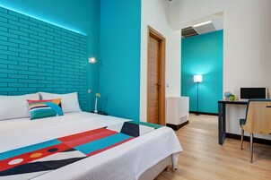A modern hotel room with a teal accent wall, a neatly made bed with a colorful geometric throw, a desk with a computer, and a bright desk lamp.