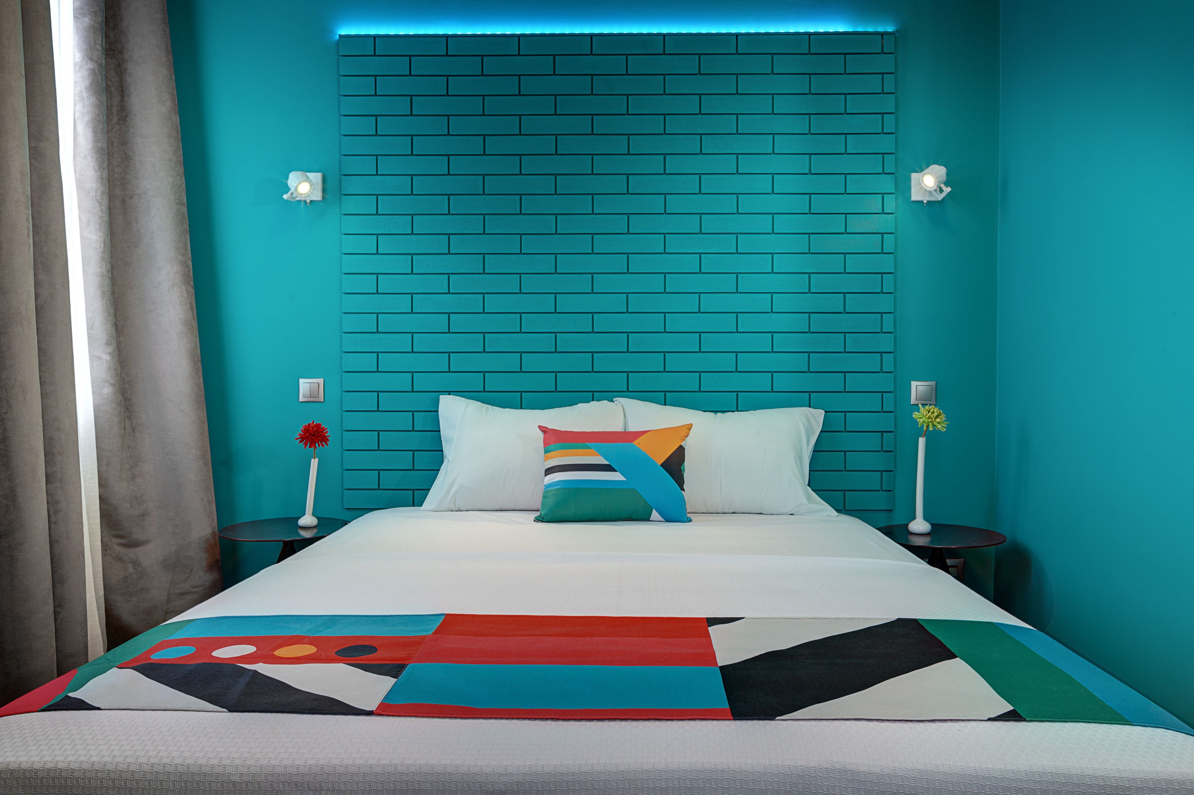 A hotel room with a teal brick headboard, white bedding, and a colorful geometric-patterned bedspread.