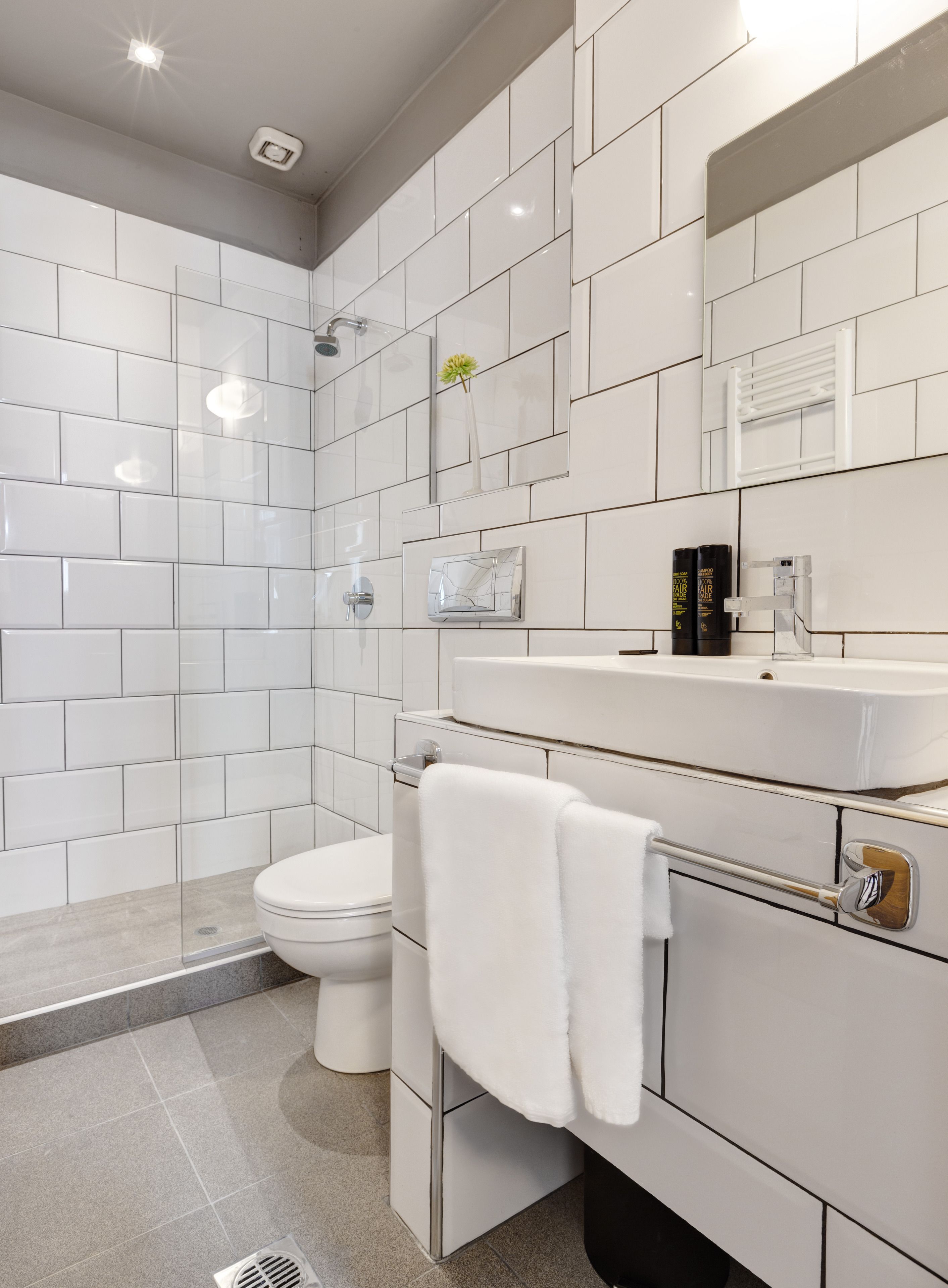 Un baño moderno con azulejos blancos, mampara de ducha de vidrio, inodoro y tocador con espejo y artículos de aseo.