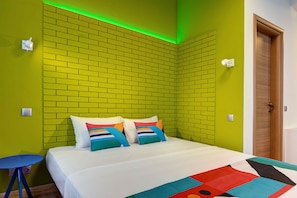 Una habitación con pared de ladrillo, una cama con almohadones de colores, una pequeña mesa azul y una puerta.