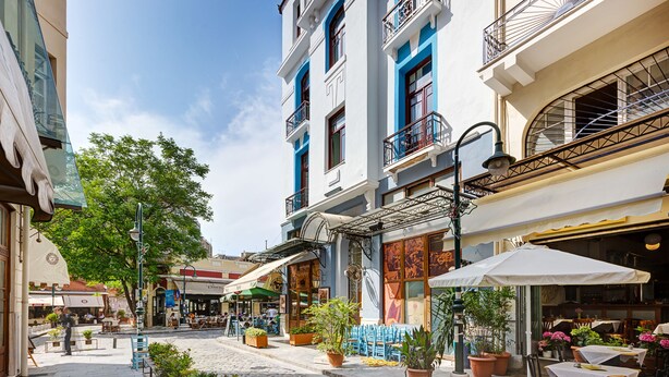 Une rue pavée bordée de terrasses, de plantes en pot et de divers bâtiments dotés de balcons et de grandes fenêtres.