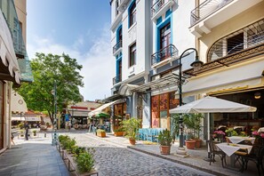 Una calle adoquinada bordeada de terrazas al aire libre, macetas con plantas y diversos edificios con balcones y grandes ventanales.