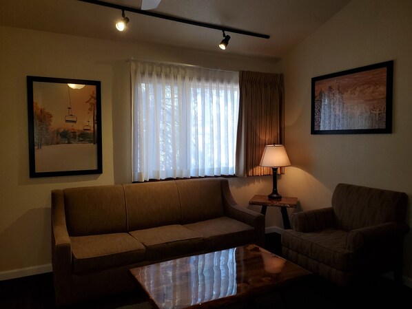 Un salón con sofá, sillón y mesa de centro. Se aprecia una ventana con cortinas translúcidas y una lámpara sobre una mesita.