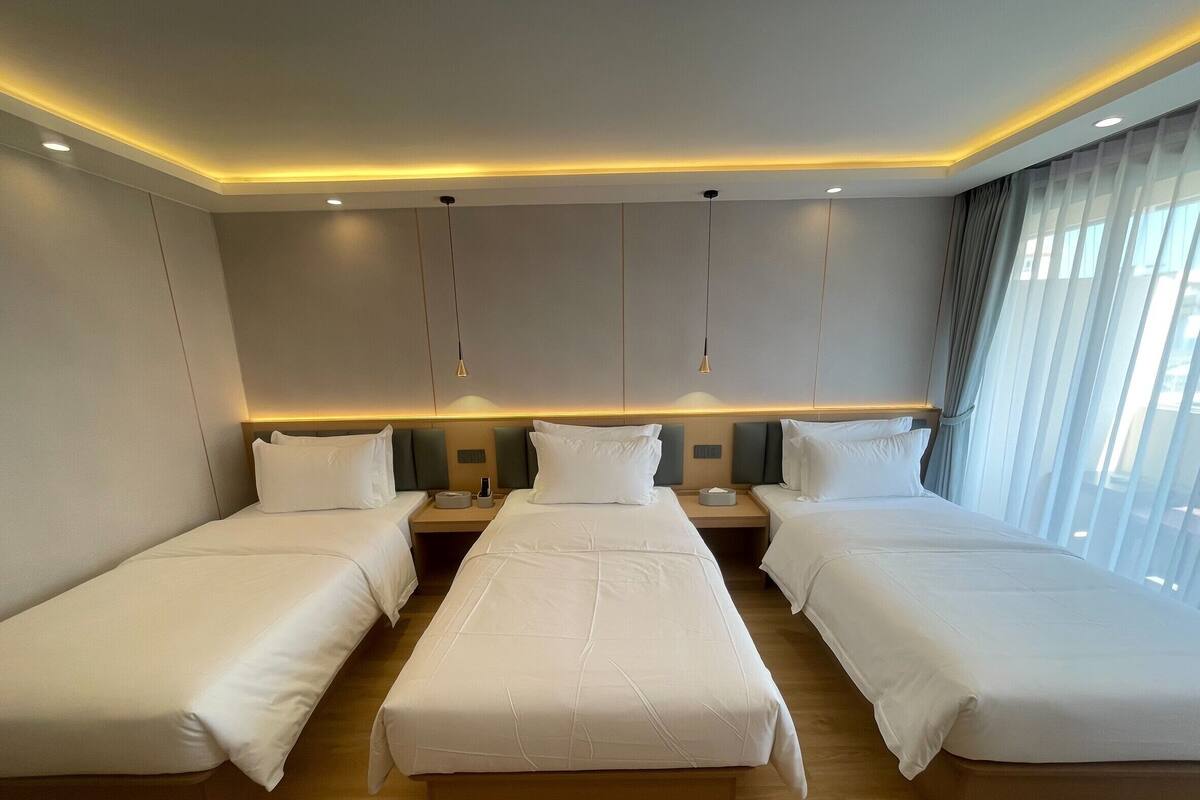 A hotel room with three beds, a wooden floor, and a large window with curtains.