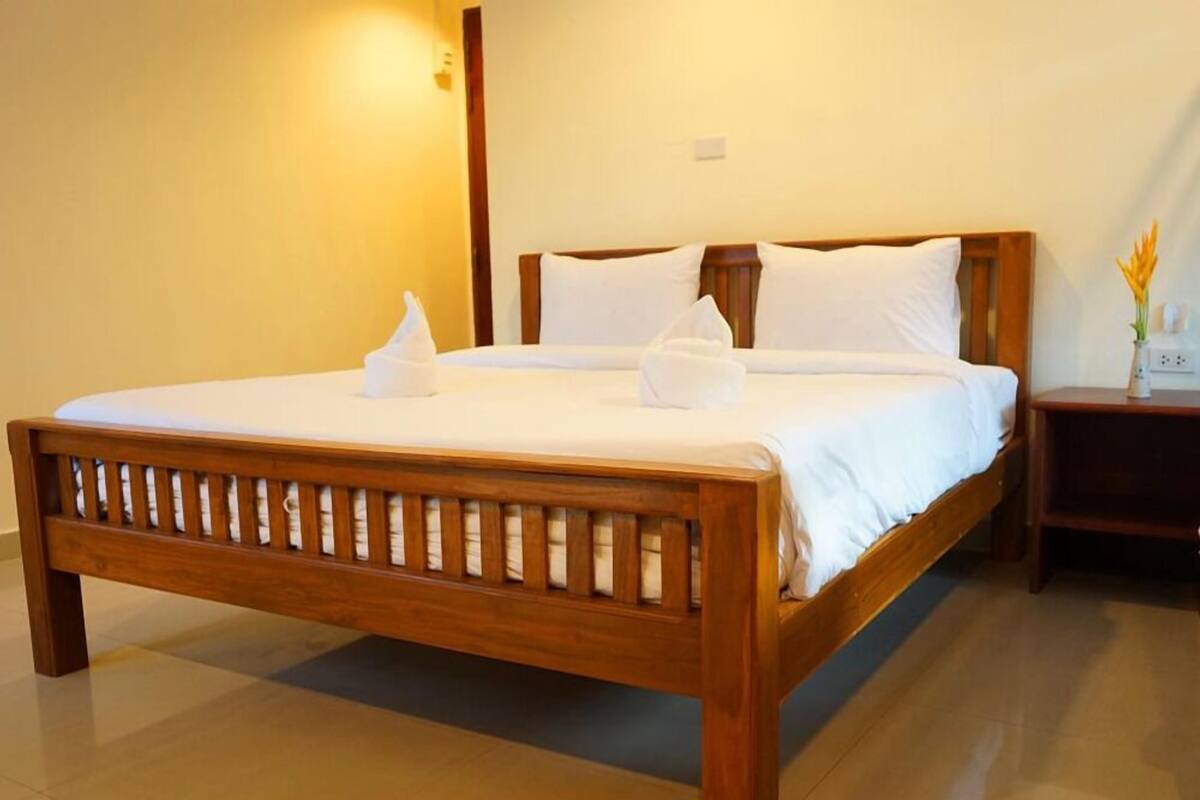 A wooden bed with white linens and pillows, a bedside table with a vase of flowers, and a wall-mounted light.