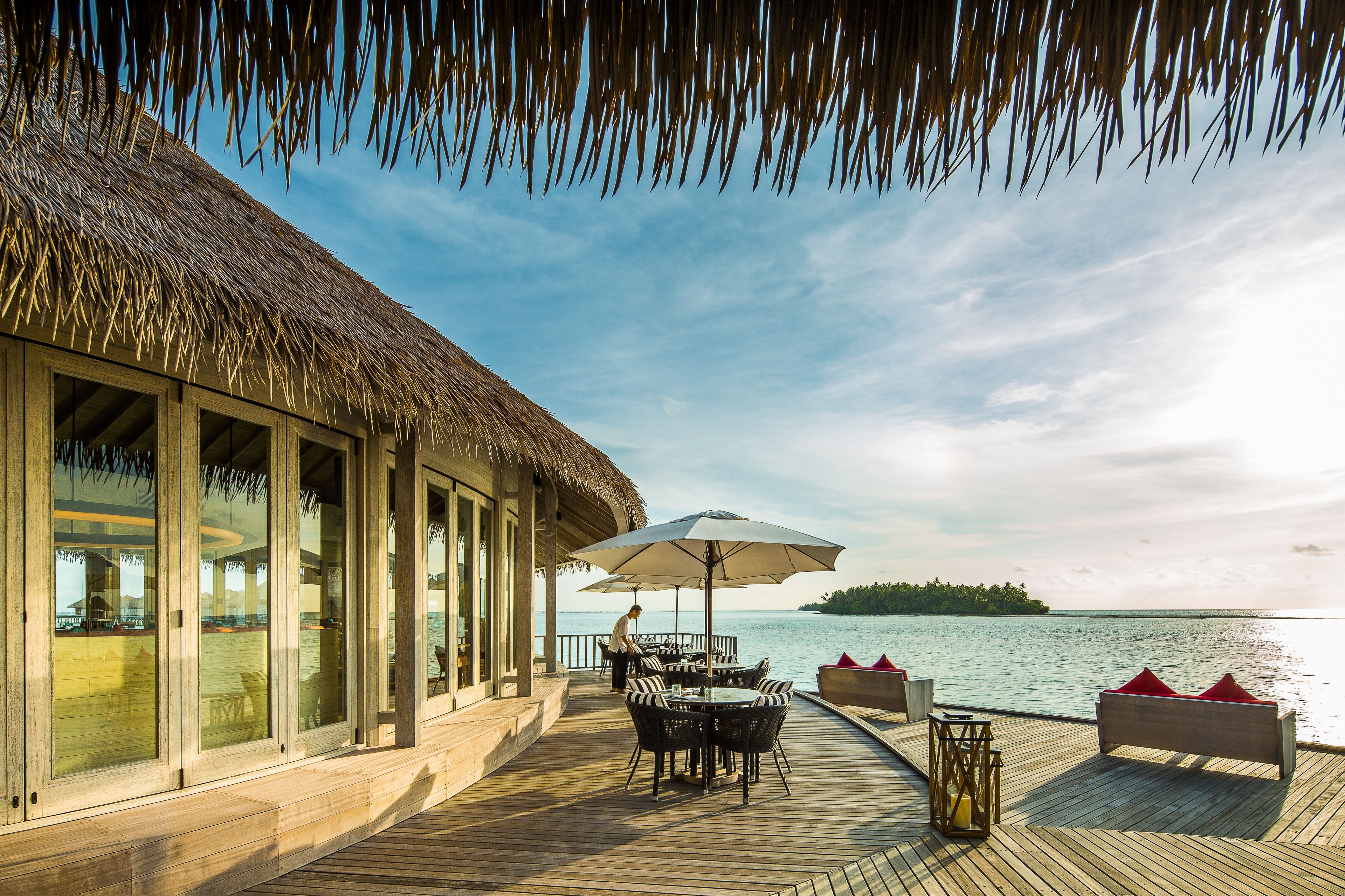 Une terrasse en bois avec des tables et des chaises, une structure surmontée d’un toit de chaume et une vue sur la mer.