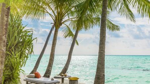 A beachside lounge with a white cushioned bench, a small round table, and a yellow cushion. - COMO Maalifushi (Maalefushi)