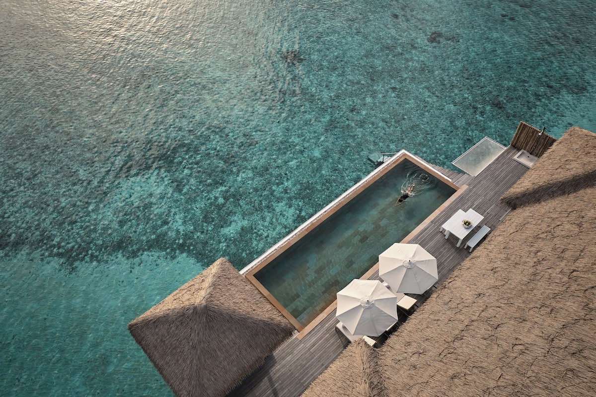 Una piscina infinita con terraza de madera y sillas reclinables blancas.