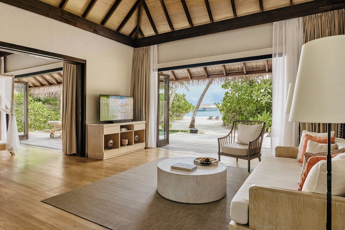 Amplia sala de estar con piso de madera, una mesa redonda para café, un sofá y un televisor sobre un mueble de madera. Grandes ventanas ofrecen vista a una playa y palmeras.