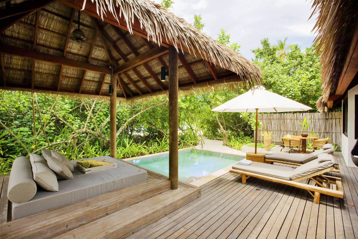 Una terraza de madera con piscina, sillas de descanso y una estructura con techo de paja.