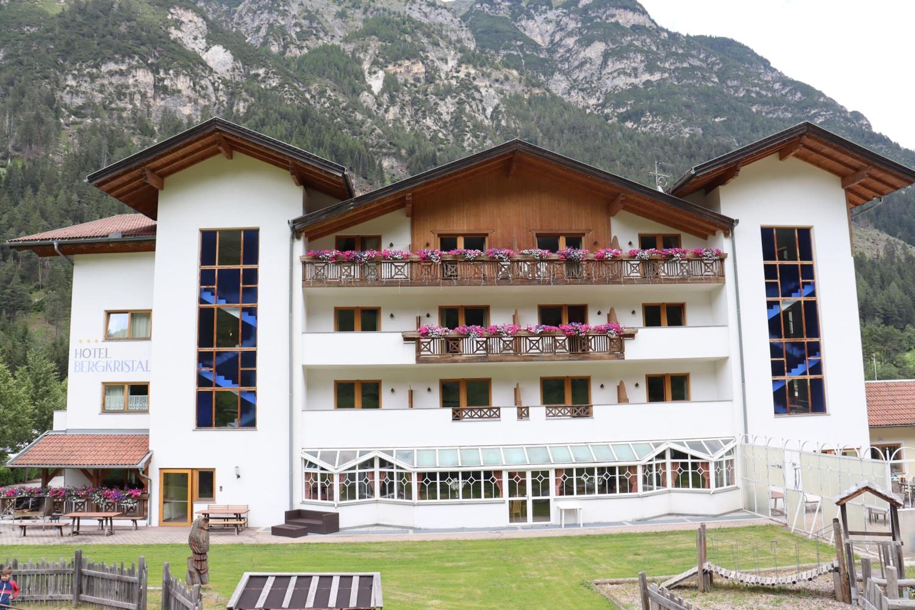 Un hotel con fachada blanca, balcones de madera con macetas de flores y montañas al fondo.