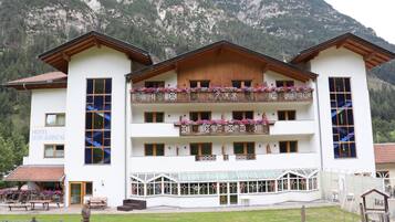 Un hotel con fachada blanca, balcones de madera con macetas de flores y montañas al fondo.