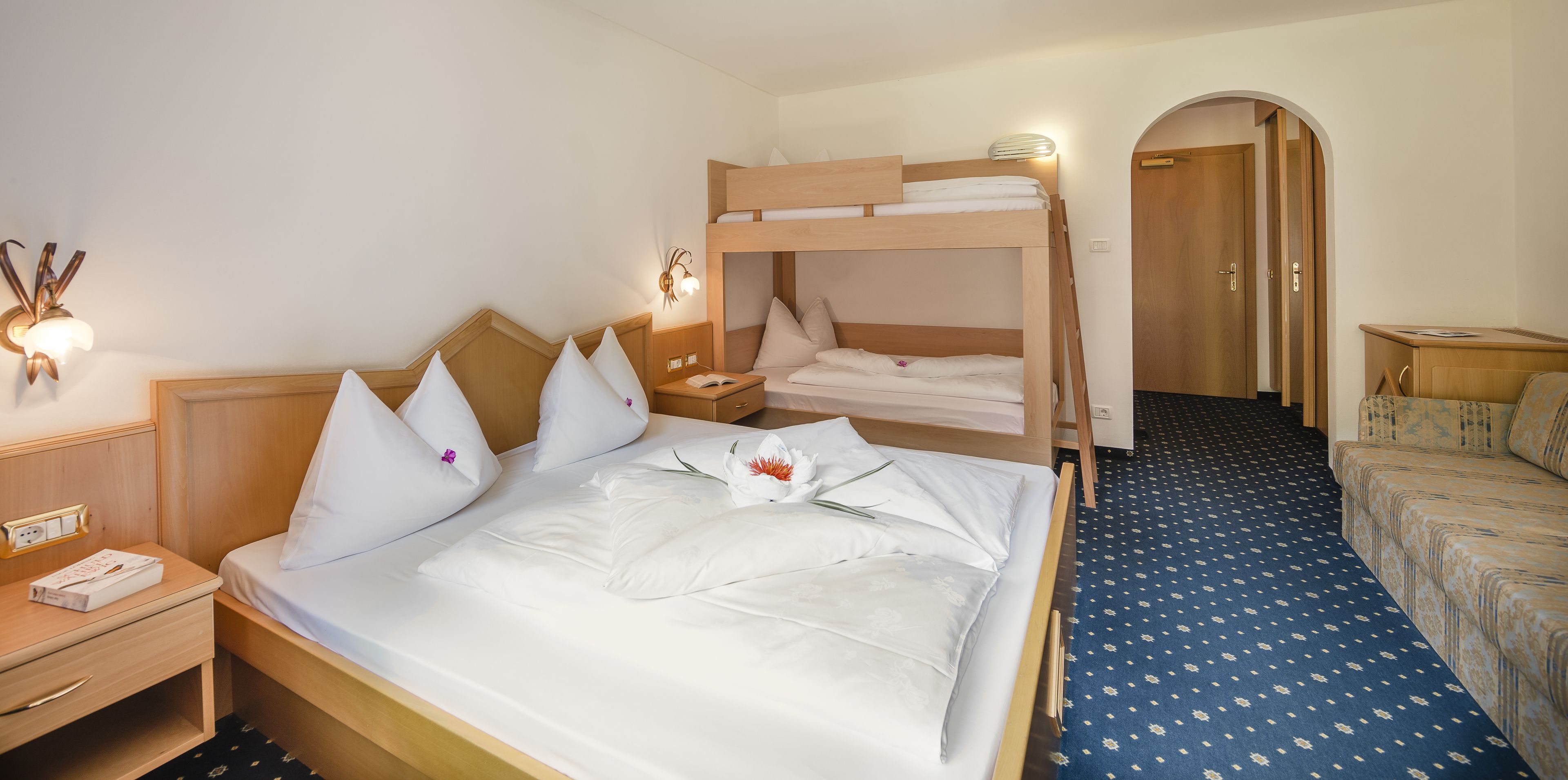 A hotel room with a bunk bed, a single bed, a desk with a lamp, and a patterned carpet.