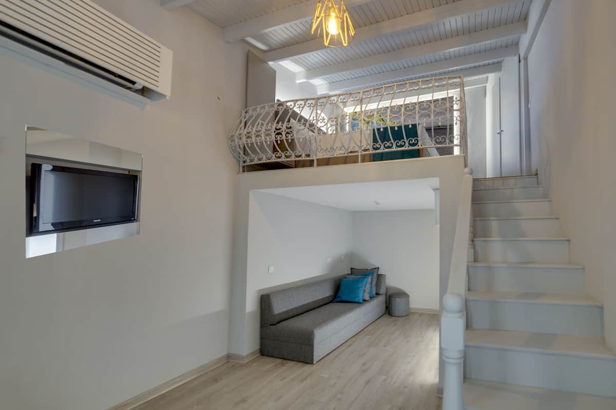 A modern living room with a grey sofa, a flat-screen TV mounted on the wall, a staircase, and a balcony with a decorative railing.